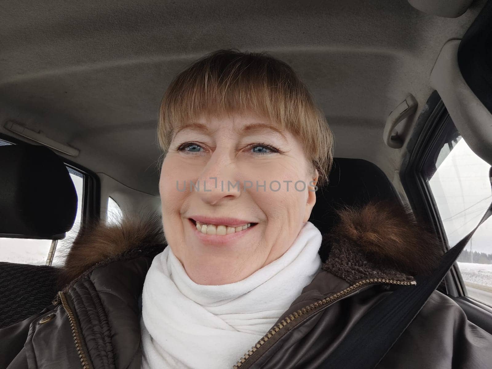 Portrait of adult cute funny blonde woman in warm clothes taking a selfie inside the car and driving it. The concept of a pleasant trip in spring, autumn, winter by keleny