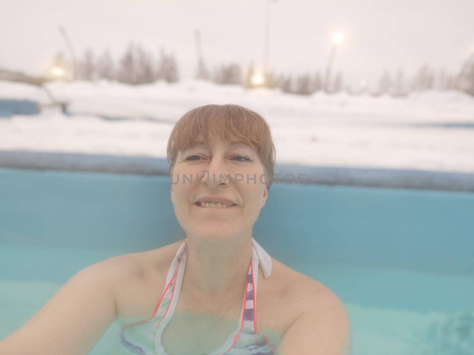 Adult mature woman takes selfie in a pool with warm hot termal mineral water in winter and white snow around. Wellness center and the concept of health care. Travel, recreation, medicine and rest