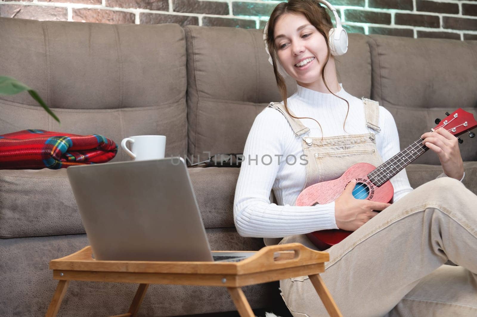 Smiling young teacher in headphones gives ukulele lesson remotely using laptop. young girl playing the ukulele sitting near the sofa at home by yanik88