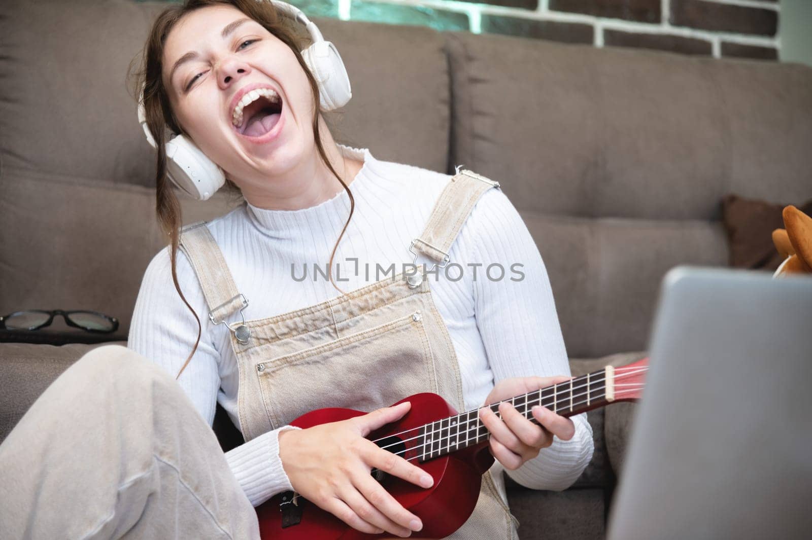 Cheerful young woman playing the ukulele, sitting in the apartment. Joyful girl holding an acoustic ukulele and singing at home. women's musical hobby, playing the ukulele by yanik88