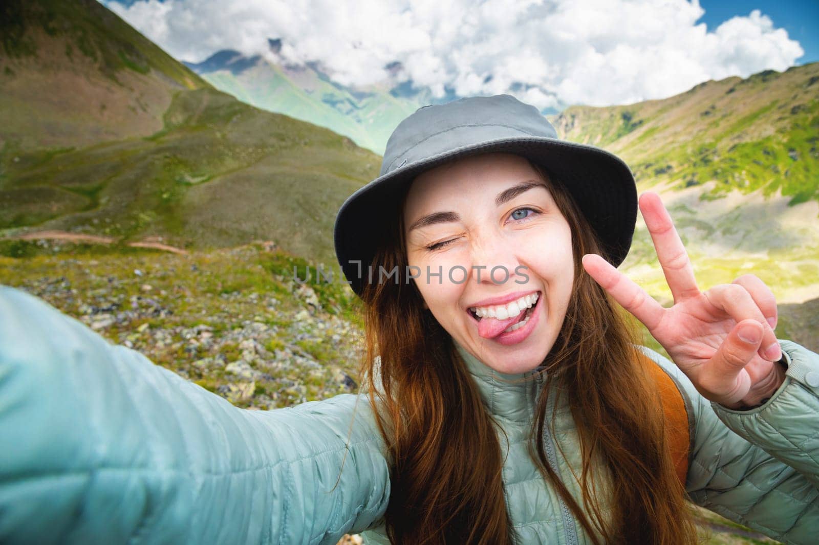 Self-portrait of a young cheerful beautiful caucasian girl with a backpack in the wild in the mountains, showing tongue, winking and smiling. Woman taking a funny selfie.