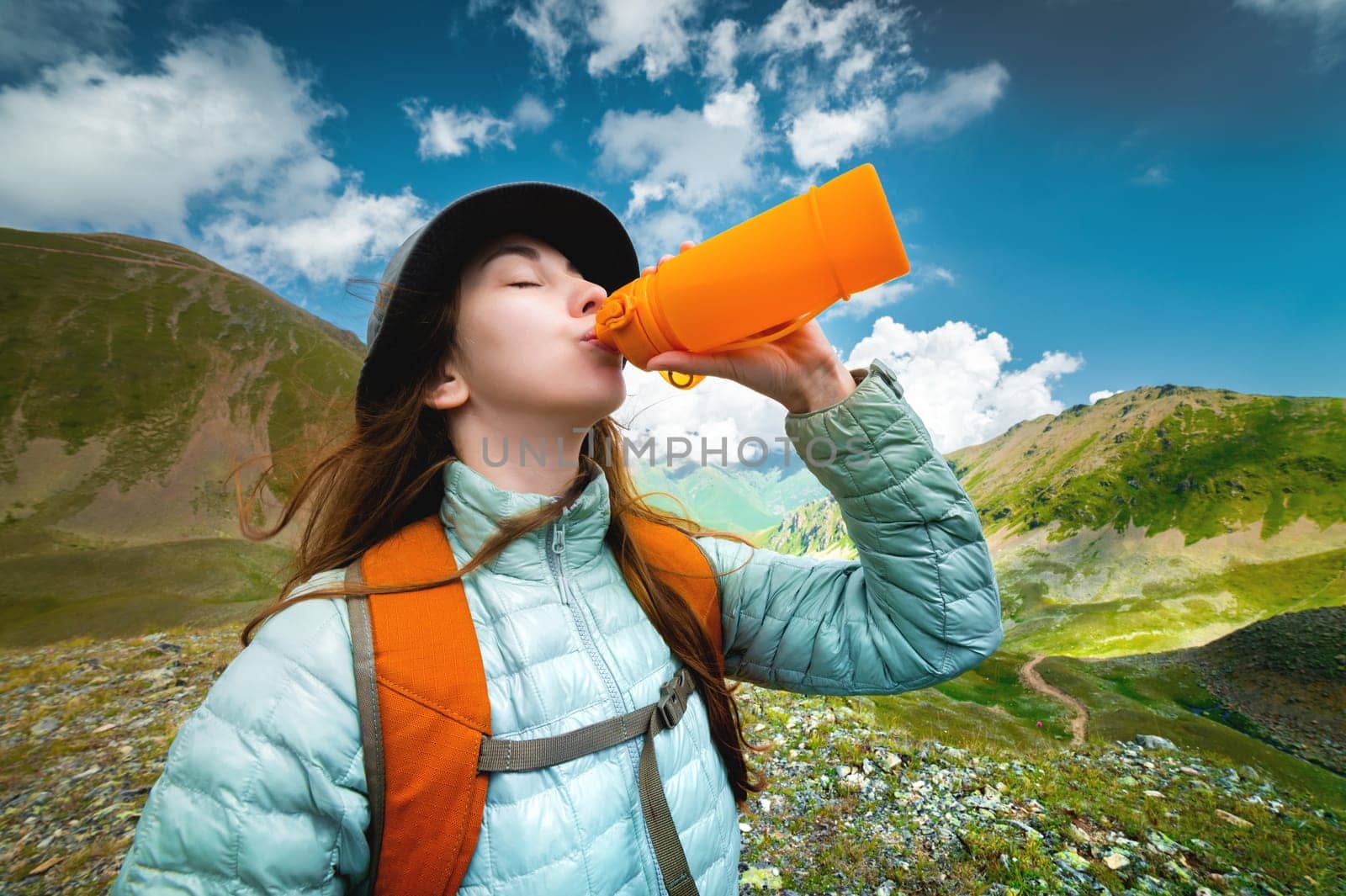 sporty caucasian female tourist drinking from water bottle in green mountains. Woman tourist with a backpack drinking from a reusable silicone water bottle in nature by yanik88