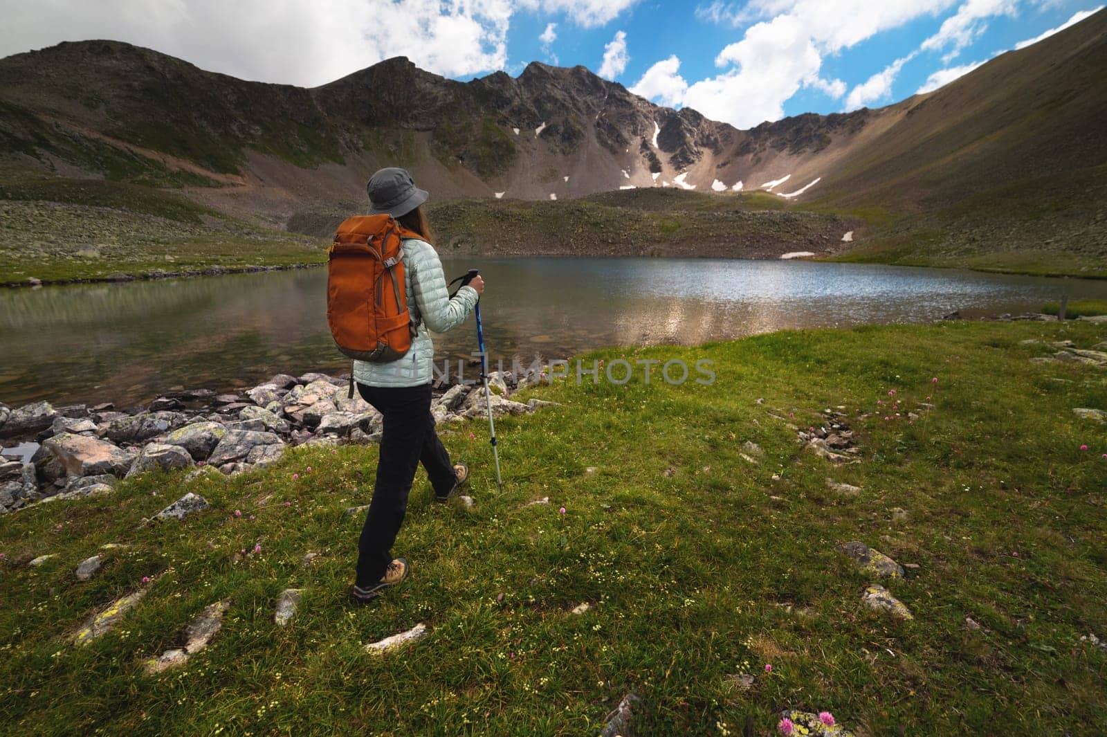 A young woman goes to a mountain lake in summer against the backdrop of picturesque mountains and a cloudy sky. Girl hiking with a backpack in the mountains.
