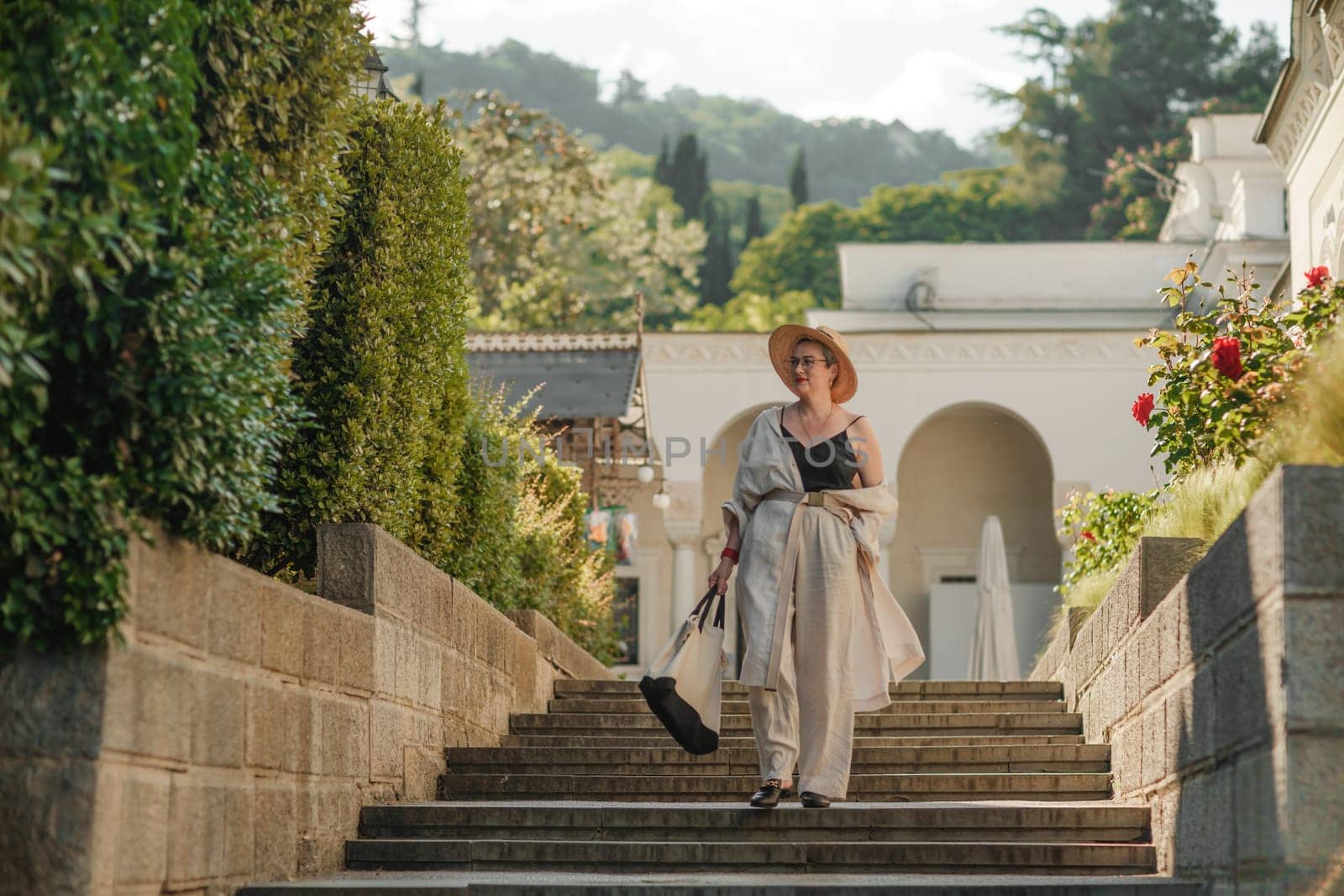 Woman on the stairs in the park. A middle-aged lady in a hat in a white outfit with a bag walks around the Livadia Palace.