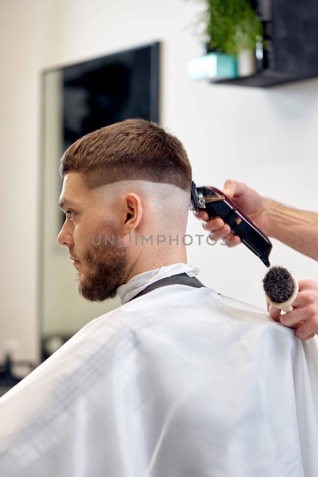Barber trim hair with clipper on handsome bearded man in barber shop.