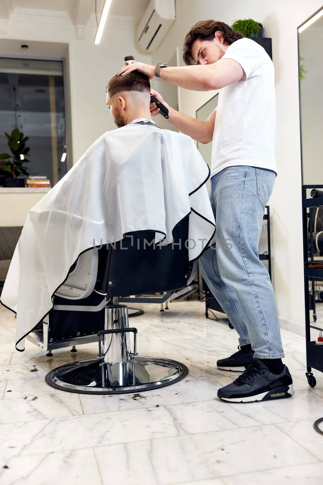 Barber trim hair with clipper on handsome bearded man in barber shop.