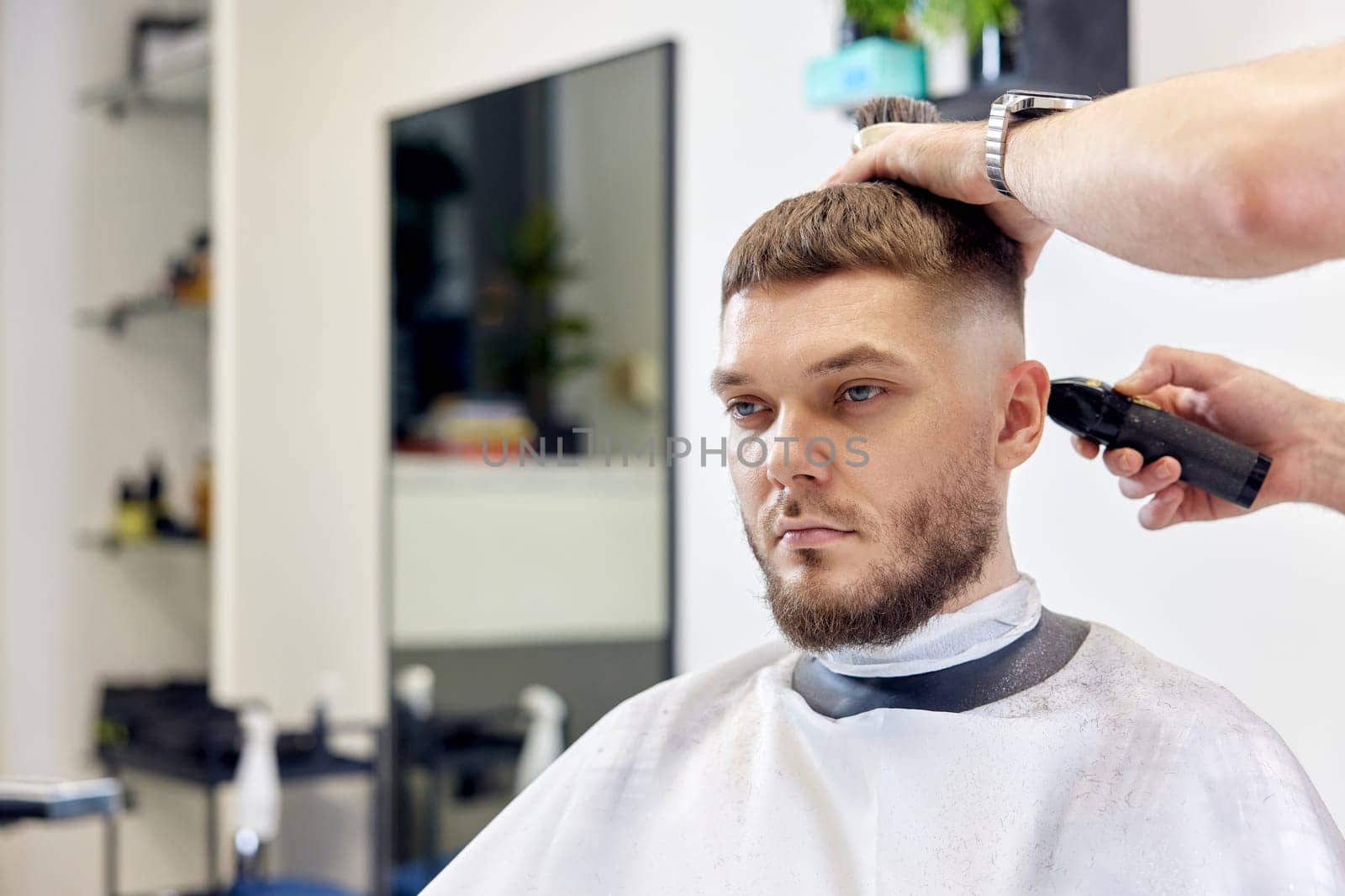 Barber trim hair with clipper on handsome bearded man in barber shop.