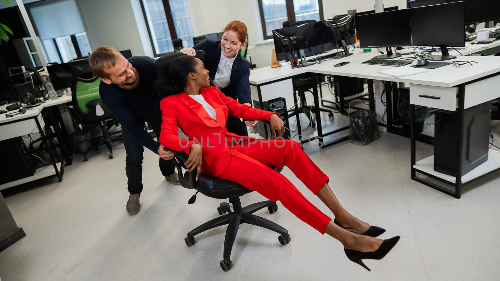 Caucasian red-haired woman, bearded caucasian man rolled African American young woman on office chair. Colleagues have fun at work