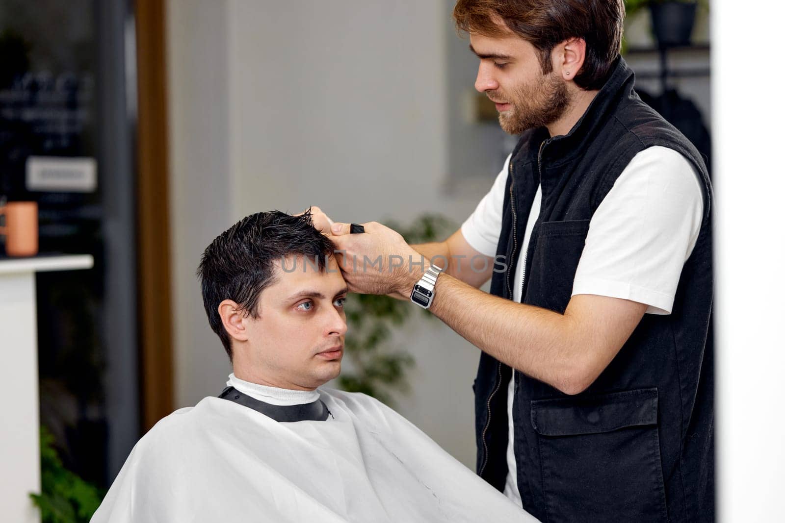professional male hairstylist combing young customer's hair in barbershop