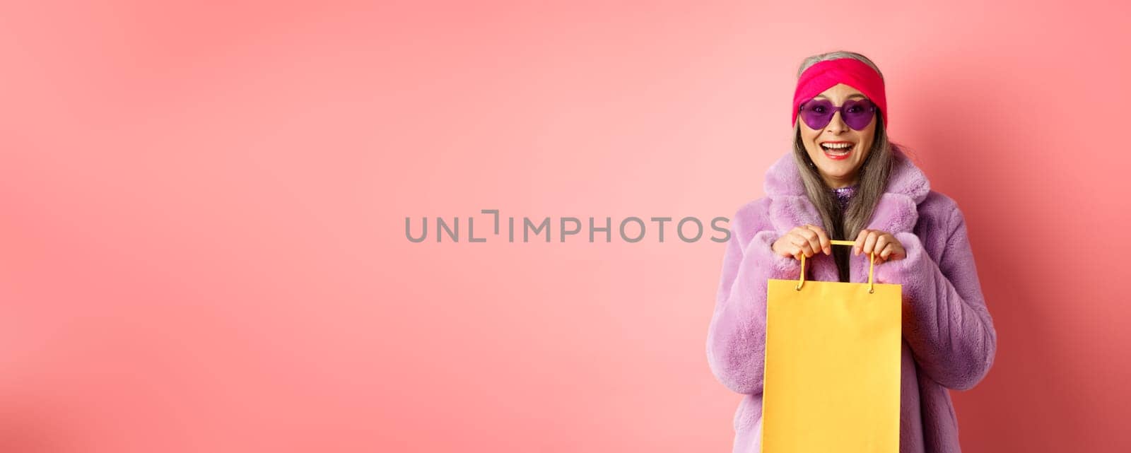 Fashionable asian senior woman going shopping in sunglasses and trendy winter clothes, holding paper bag from store and smiling happy at camera, pink background.