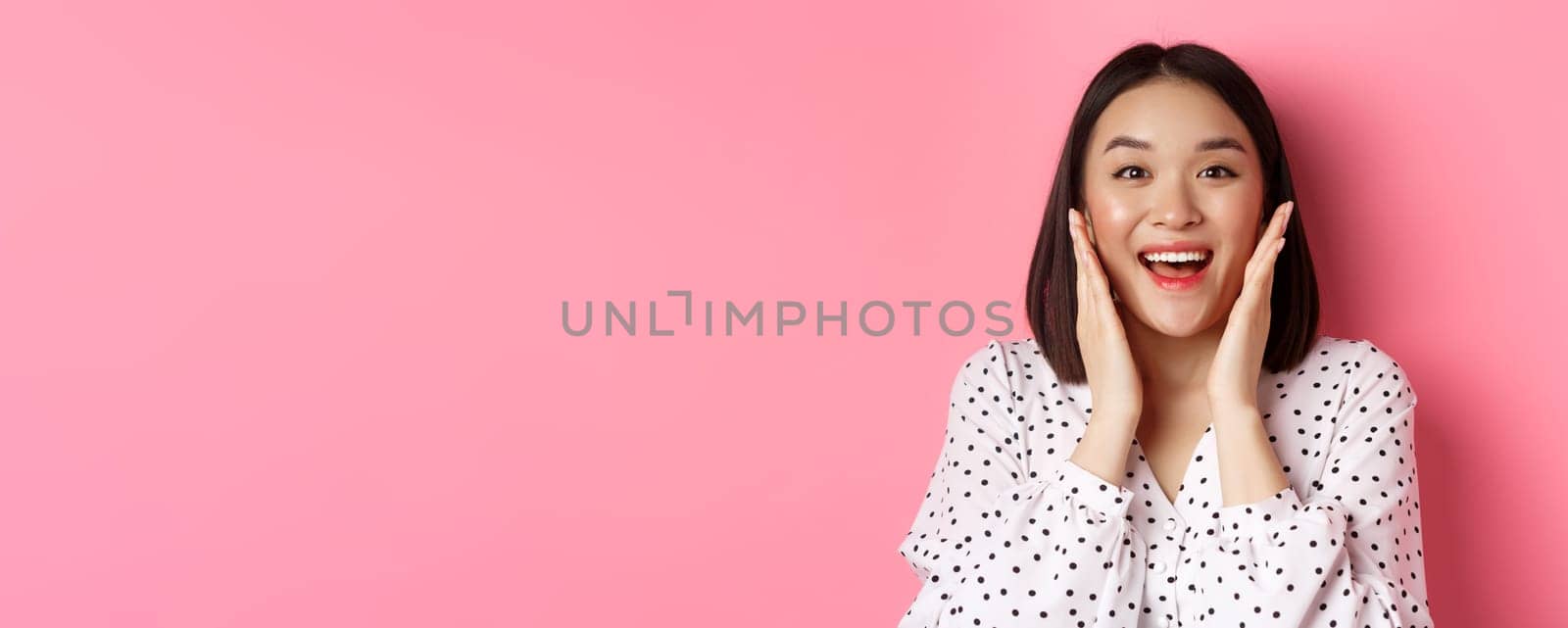 Close-up of beautiful asian woman touching clean skin face and staring at camera amazed, standing over pink background.