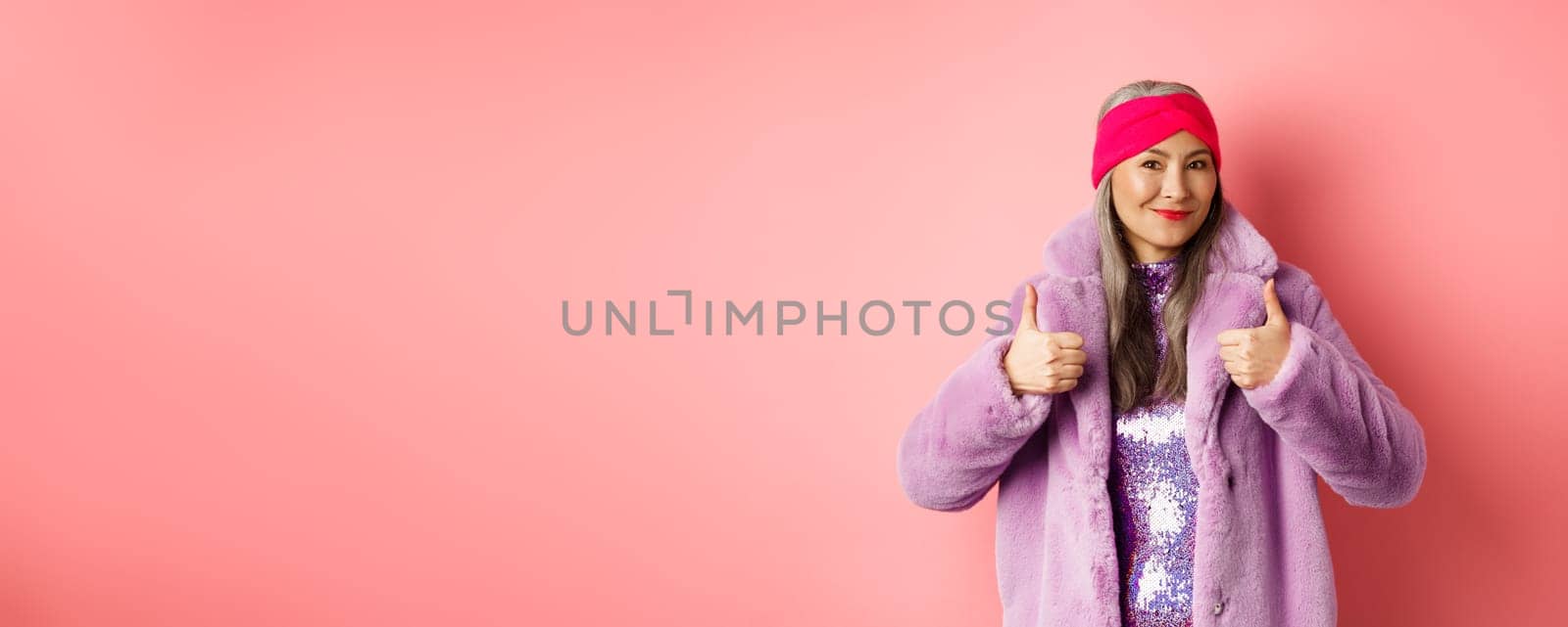 Fashion and shopping concept. Asian fashionable lady in winter coat showing thumbs-up, smiling and looking satisfied, recommending company promo, pink background.