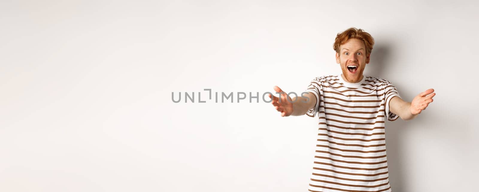 Cheerful man with red curly hair reaching hands forward, stretch out arms to welcome or congratulate you, smiling happy, standing over white background.