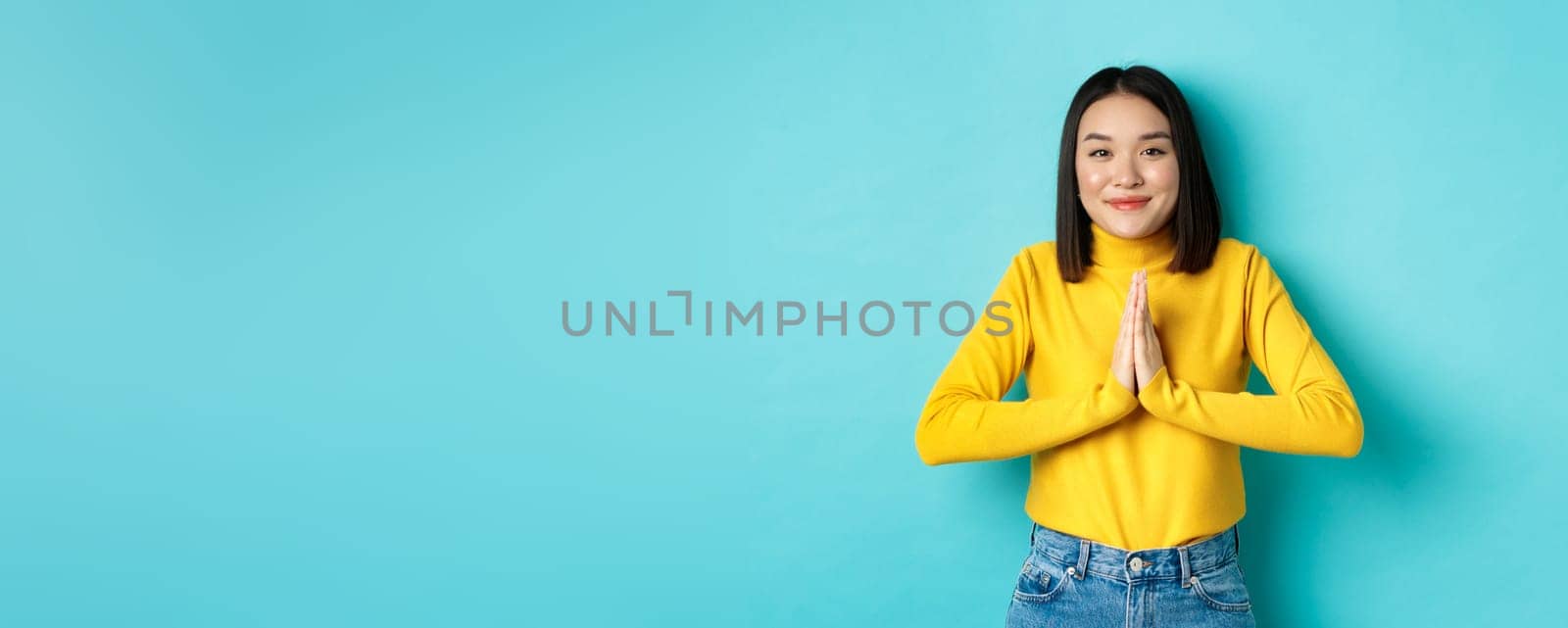 Cute asian woman in trendy outfit saying thank you, holding hands in namaste, begging gesture, smiling grateful at camera, standing over blue background.