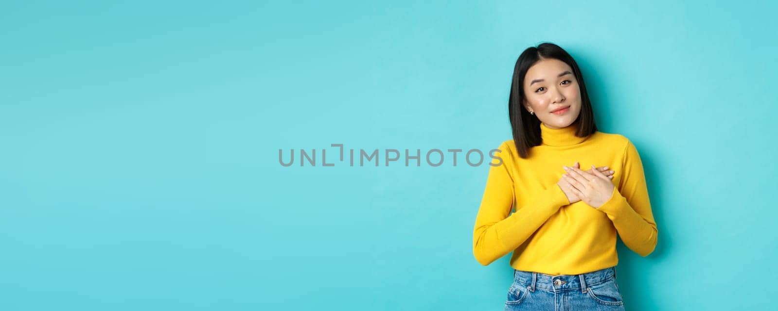 Portrait of beautiful heartfelt woman holding hands on heart, smiling and listening compassionate, standing over blue background in yellow pullover.