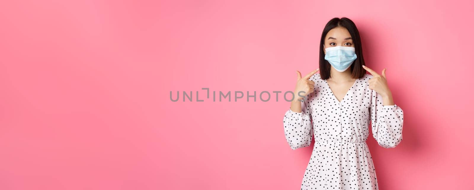 Coronavirus, social distancing and lifestyle concept. Cute asian woman pointing at face mask, asking to use measures against covid-19, standing over pink background.