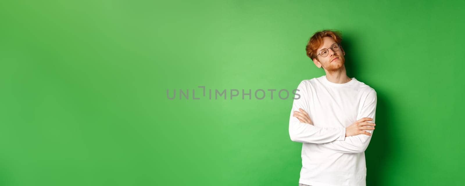 Confident young man with red hair, wearing glasses, looking smug at right side copy space, cross arms on chest self-assured, standing over green background.