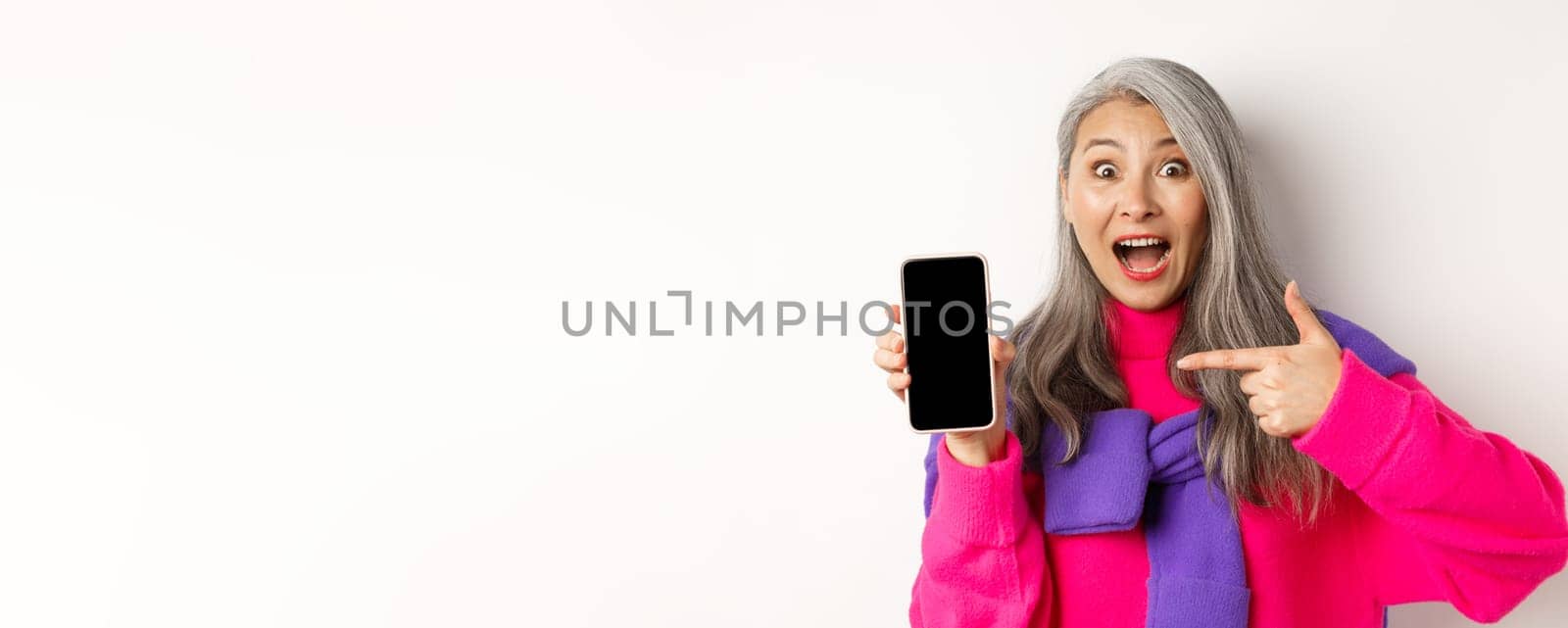 Online shopping. Beautiful asian grandmother smiling, pointing finger at smartphone blank screen, looking amazed, showing mobile application, standing over white background.