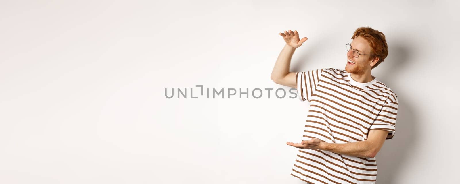 Happy and satisfied caucasian redhead man introduce something big, showing large product and smiling pleased, white background.