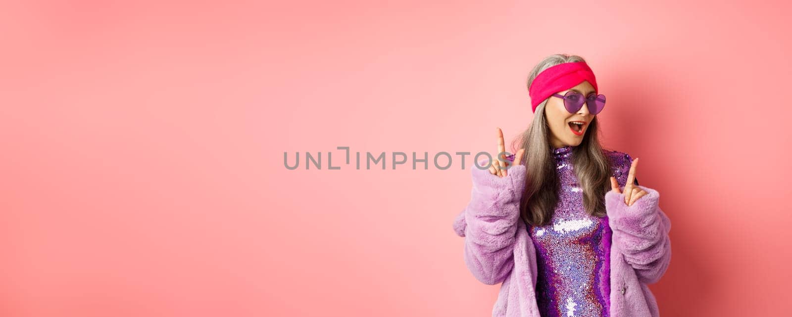 Hipster granny in sunglasses and glittering dress posing for photo with peace, victory signs, standing sassy against pink background.