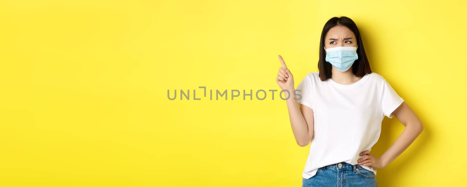 Covid, health care and pandemic concept. Asian woman in medical mask and white t-shirt pointing finger at upper left corner logo, showing promotion, yellow background.