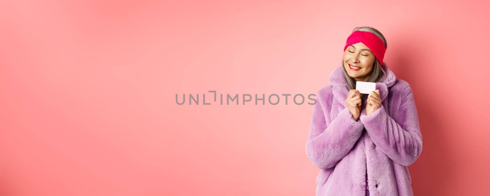 Shopping and fashion concept. Fashionable asian woman in purple faux fur coat, looking happy and showing plastic credit card, smiling cheerful, standing over pink background.