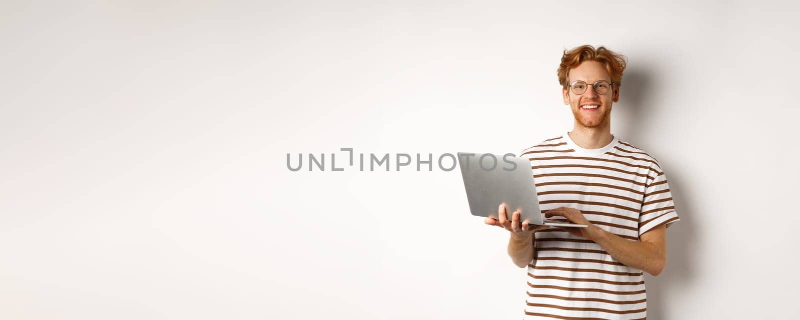 Smiling young man working on laptop and looking joyful, standing over white background.