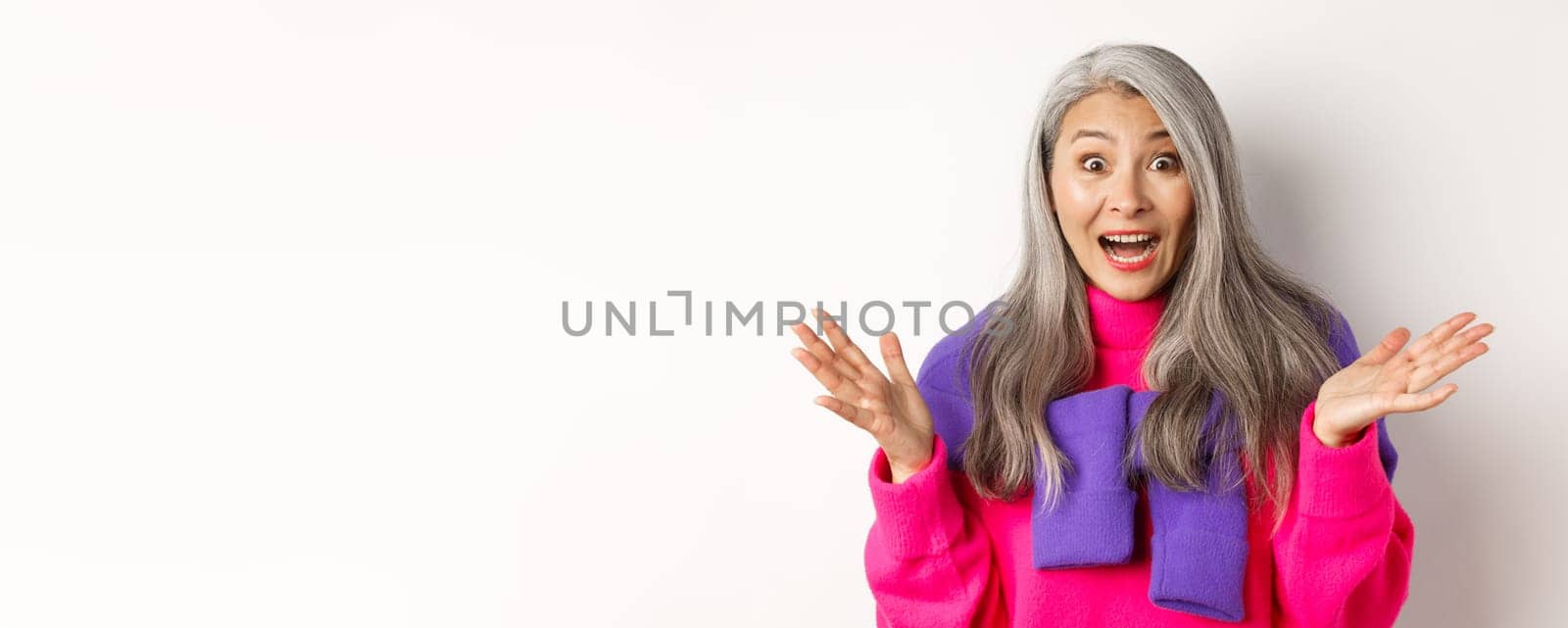 Close-up of excited asian senior woman spread hands sideways, gasping and looking amazed and surprised, hear great news, standing over white background.