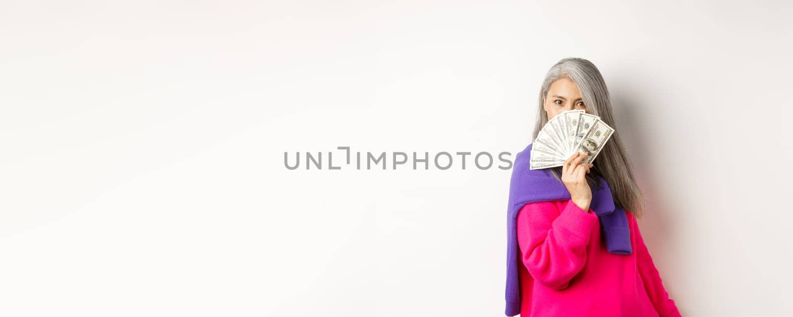 Shopping concept. Rich and fashionable korean senior woman hiding face behind money, looking at camera, standing over white background by Benzoix