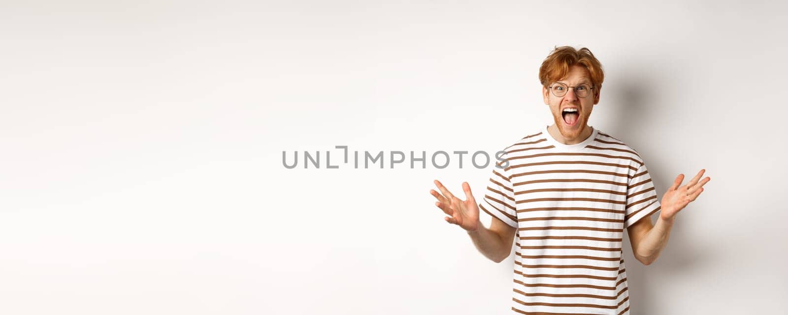 Frustrated man with red hair shaking hands and yelling at camera, looking angry and displeased, standing over white background.