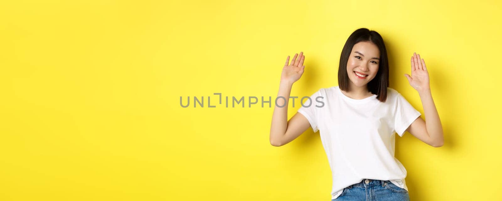 Friendly young asian woman saying hello, raising empty hands up and smiling, greeting you, standing over yellow background by Benzoix