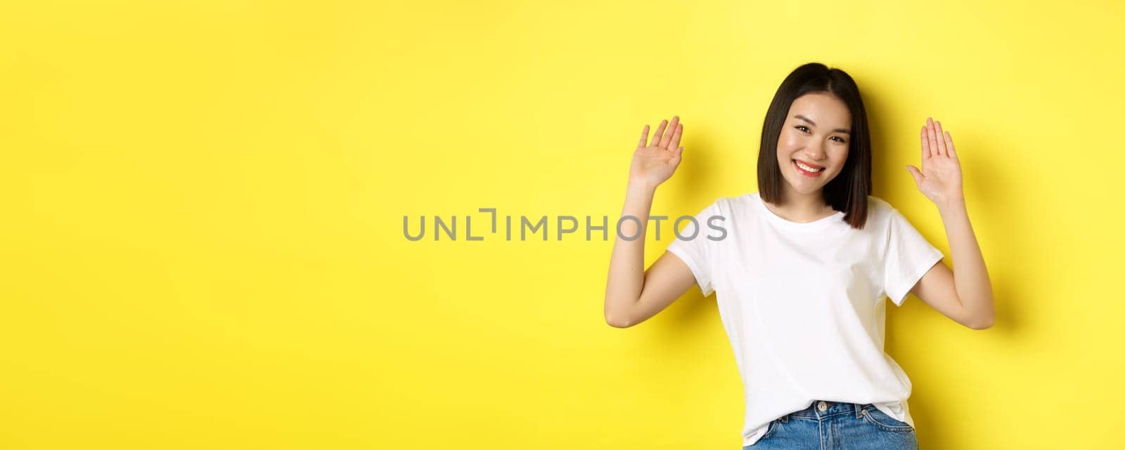 Friendly young asian woman saying hello, raising empty hands up and smiling, greeting you, standing over yellow background.