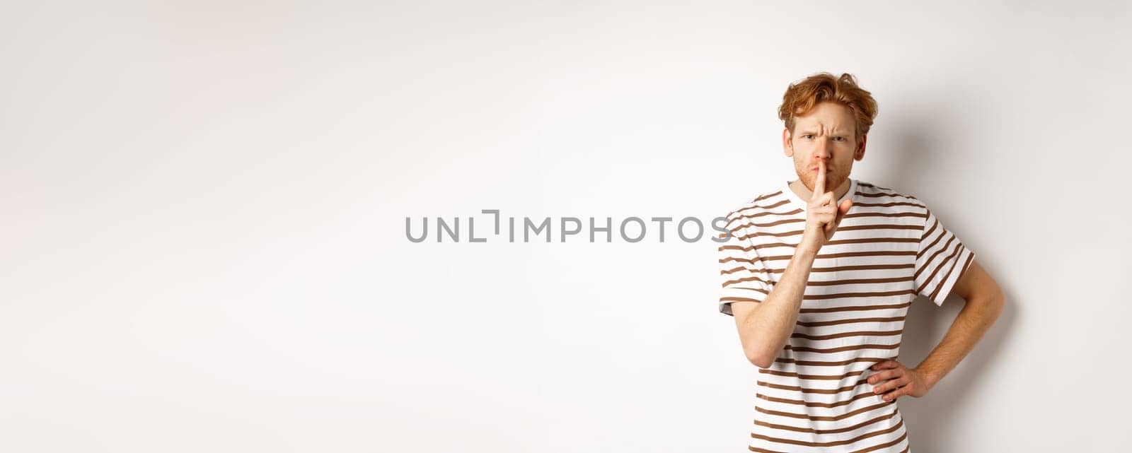 Angry redhead man looking displeased and hushing at camera, making shh gesture, standing over white background.