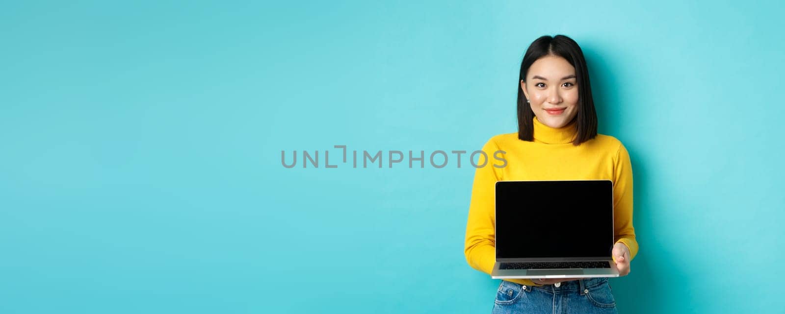 Beautiful and stylish asian woman demonstrate product on screen, showing empty laptop display and smiling, standing over blue background by Benzoix