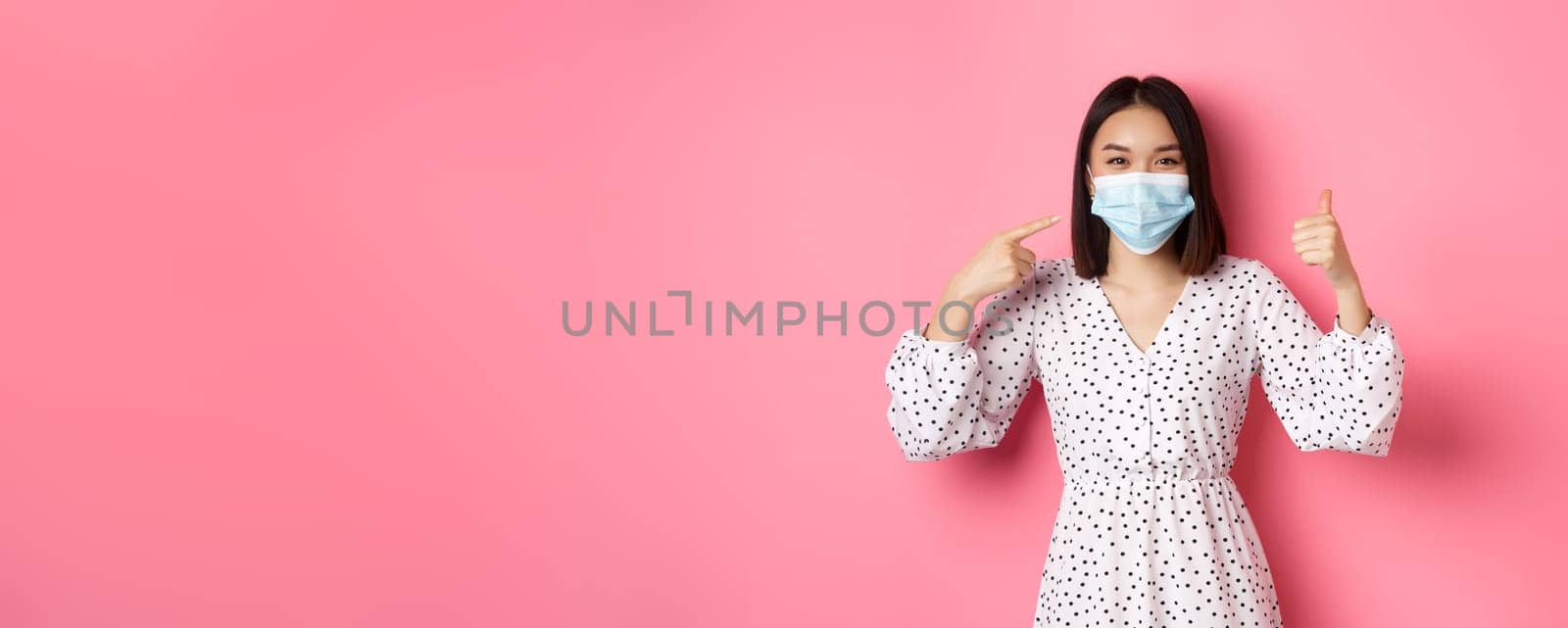 Coronavirus, social distancing and lifestyle concept. Cute asian woman pointing at face mask, showing thumb-up, support using preventive measures from covid-19, pink background.