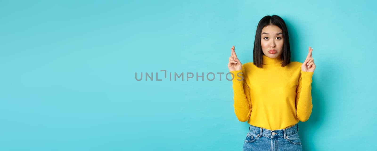 Silly hopeful asian girl making wish, pucker lips and looking at camera with dreamy glance, cross fingers for good luck, standing over blue background.