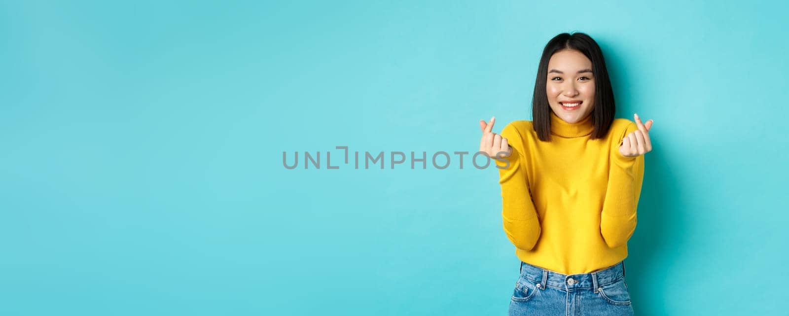 Beautiful asian girl in yellow sweater, smiling and showing finger hearts, standing happy against blue background by Benzoix