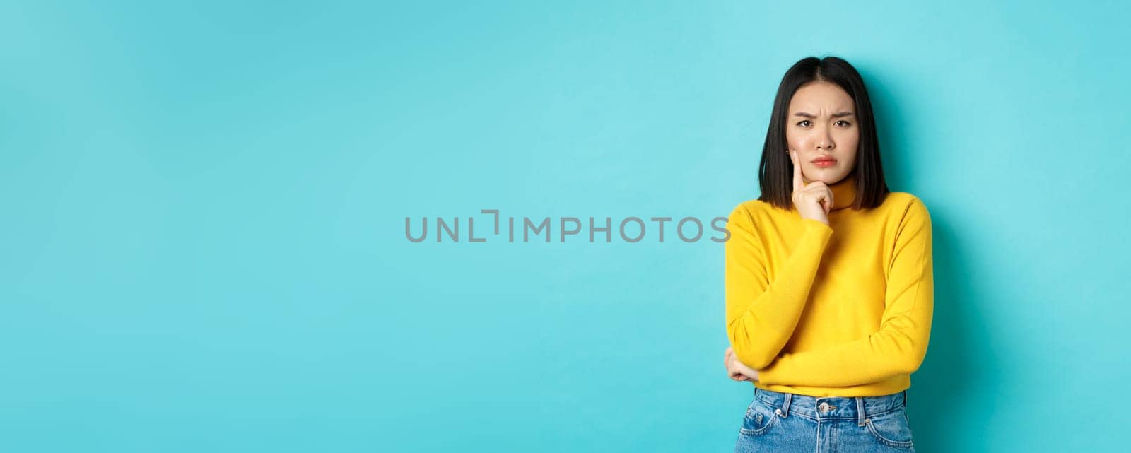 Image of pensive and serious asian woman touching chin, frowning and staring at camera puzzled, making difficult choice, standing over blue background.