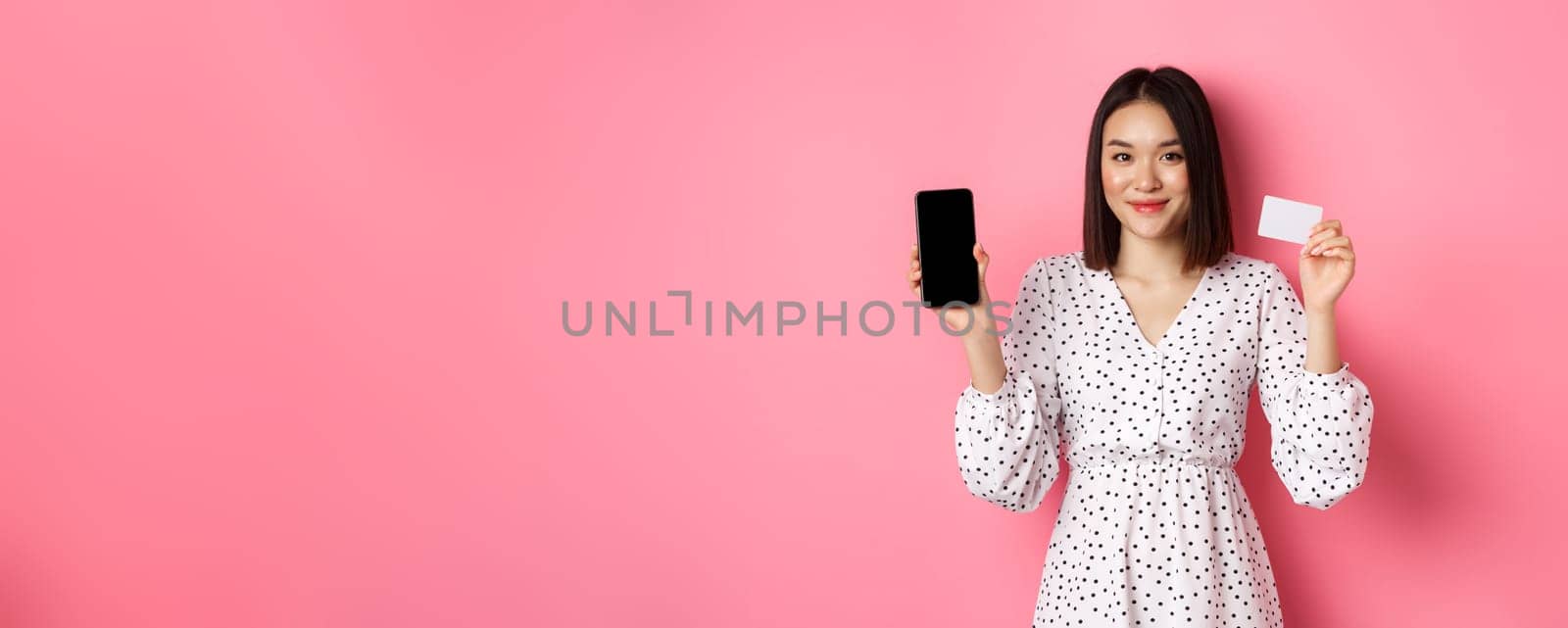 Cute asian woman shopping online, showing bank credit card and mobile screen, smiling and looking at camera, standing over pink background.