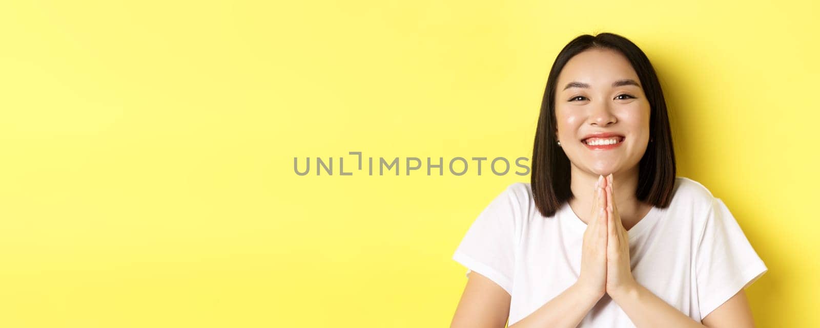 Close up of cute asian woman saying thank you and smiling, holding hands in namaste, pray gesture, standing over yellow background.