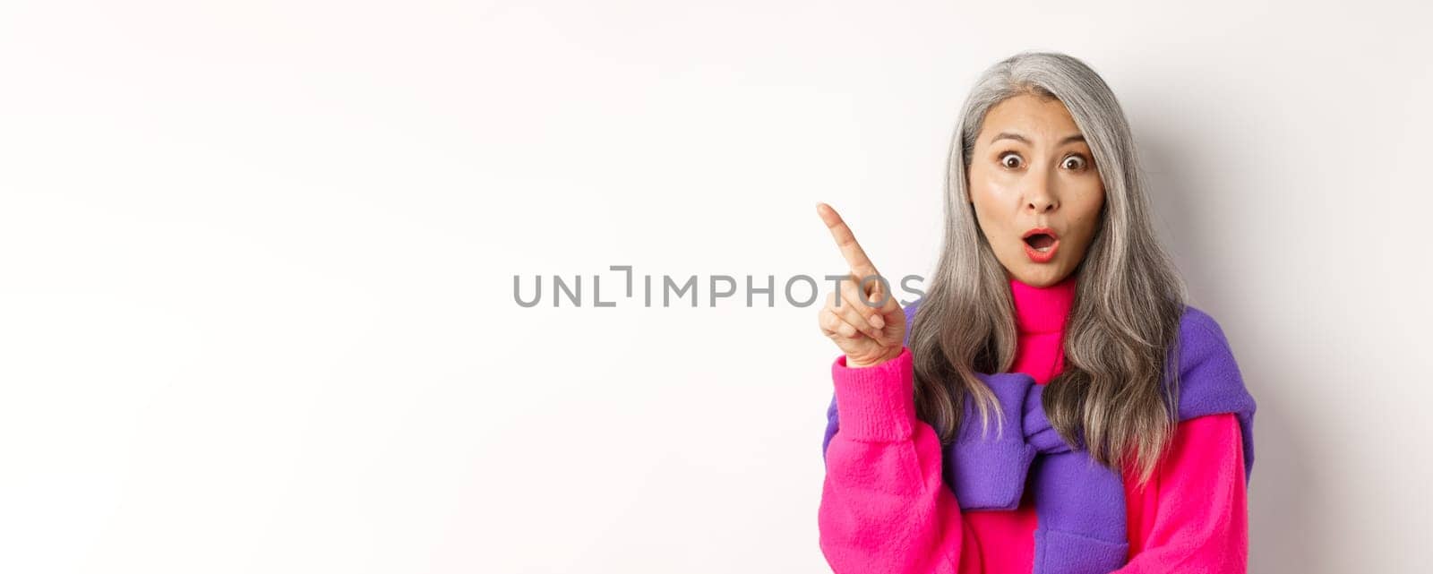 Fashion. Close up of amazed asian senior woman pointing finger upper left corner, gasping and looking impressed at camera, standing over white background.