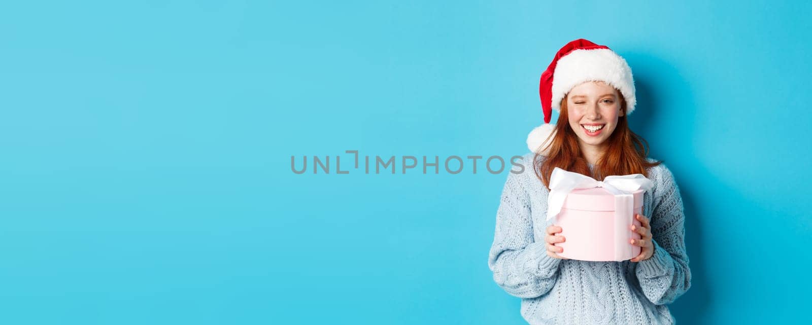 Winter holidays and Christmas Eve concept. Cute redhead girl in sweater and Santa hat, holding New Year gift and looking at camera, standing against blue background.