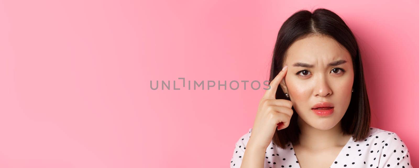 Beauty and skin care concept. Headshot of cute asian woman staring confused, pointing finger at head at frowning, standing over pink background.