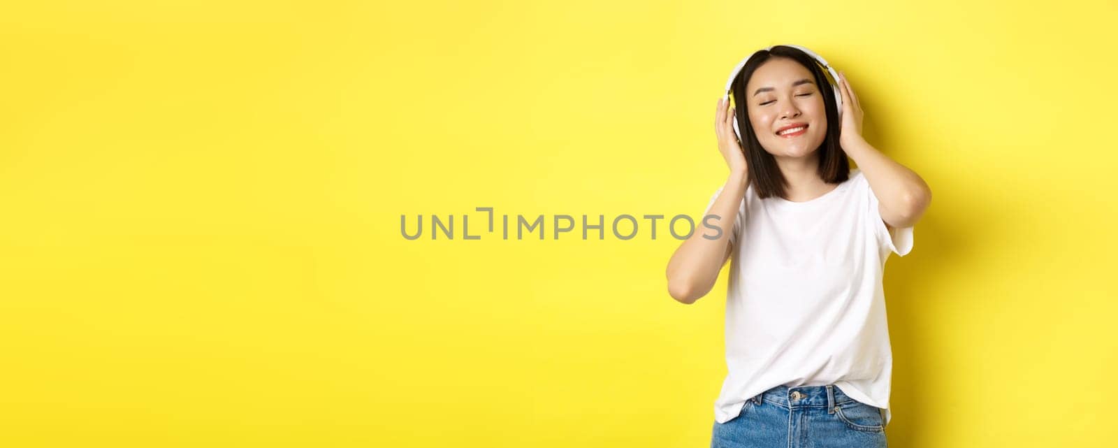 Attractive asian woman enjoying listening music in wireless headphones, smiling pleased and dancing with eyes closed, standing over yellow background by Benzoix