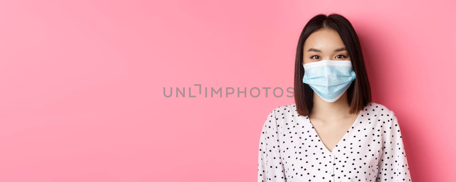 Covid-19, pandemic and lifestyle concept. Close-up of beautiful asian woman in face mask smiling with eyes, wearing protection from coronavirus in public, pink background by Benzoix