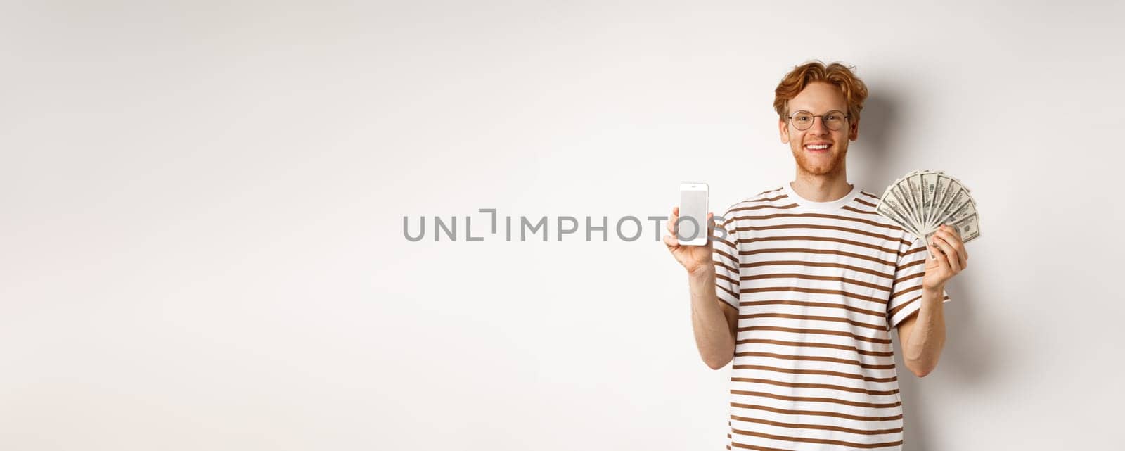 Smiling young redhead man in glasses showing smartphone blank screen and money, standing over white background by Benzoix