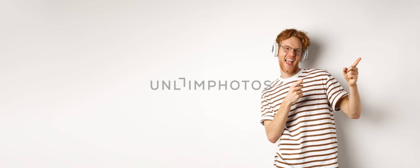 Technology concept. Happy redhead man listening music in headphones and dancing and pointing fingers right, standing over white background.