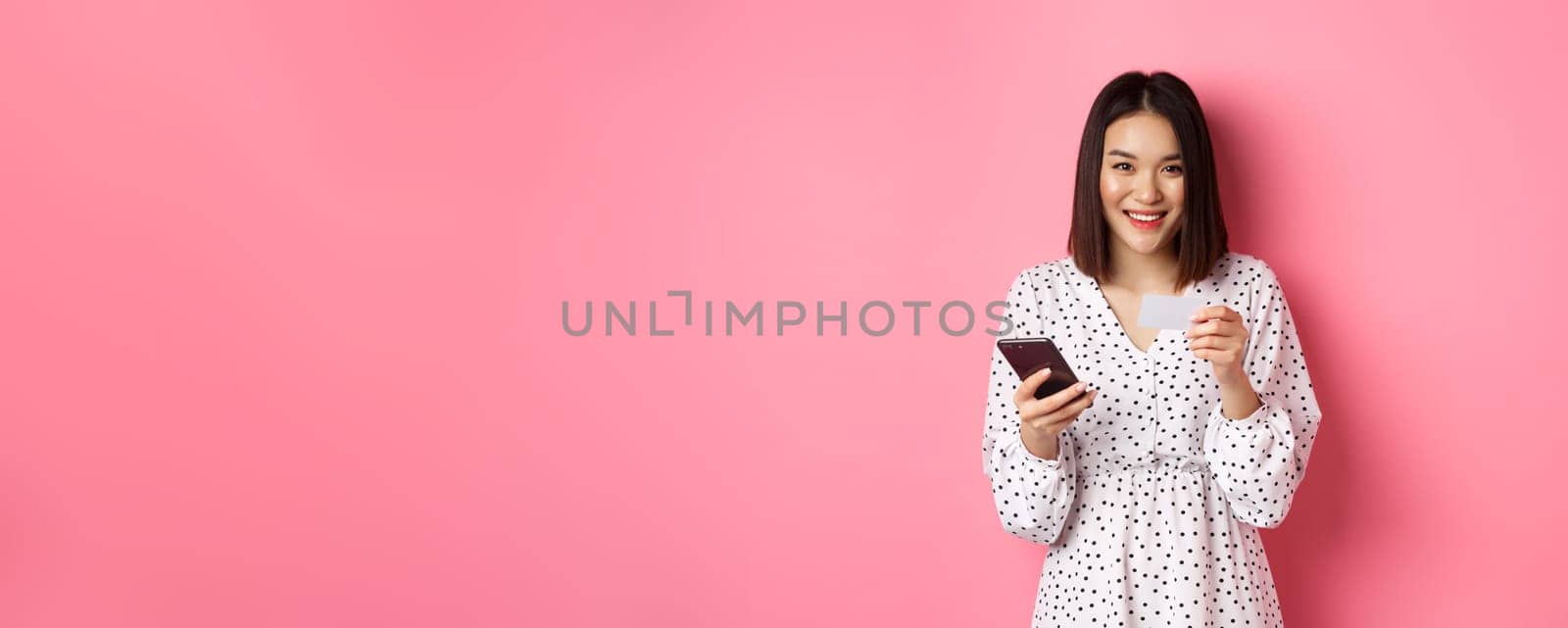 Beautiful asian woman enter credit card info in mobile phone app, shopping online, paying for order on smartphone, standing over pink background by Benzoix