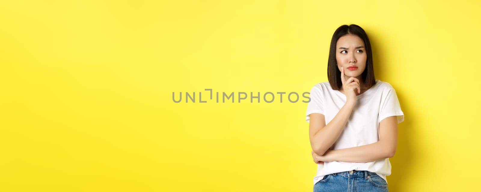 Beauty and fashion concept. Pensive asian woman thinking, looking thoughtful while pondering something, standing over yellow background.