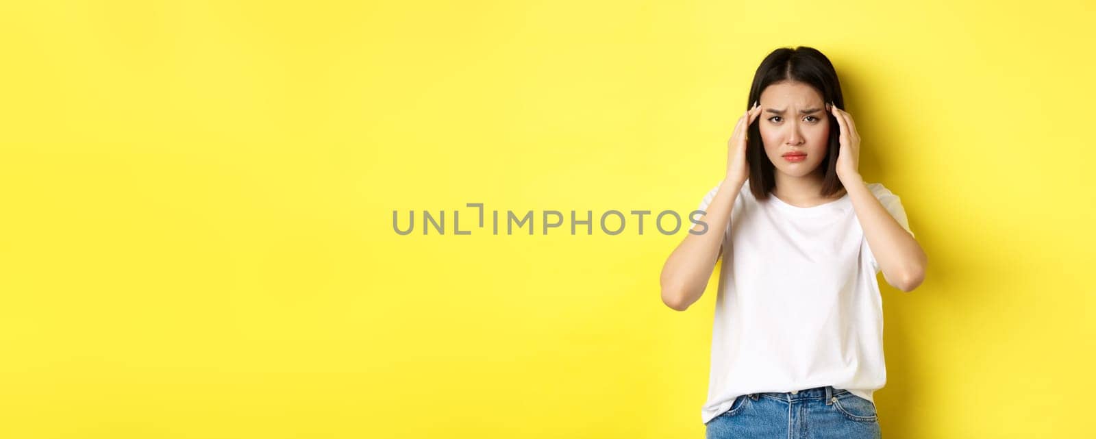 Sad asian girl touching head and frowning, feeling sick, having headache, standing in white t-shirt over yellow background.